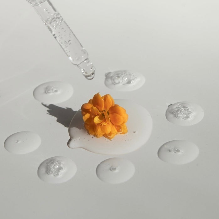 Pipette and liquid serum forming loose, transparent circles on a white background with n orange flower in the middle